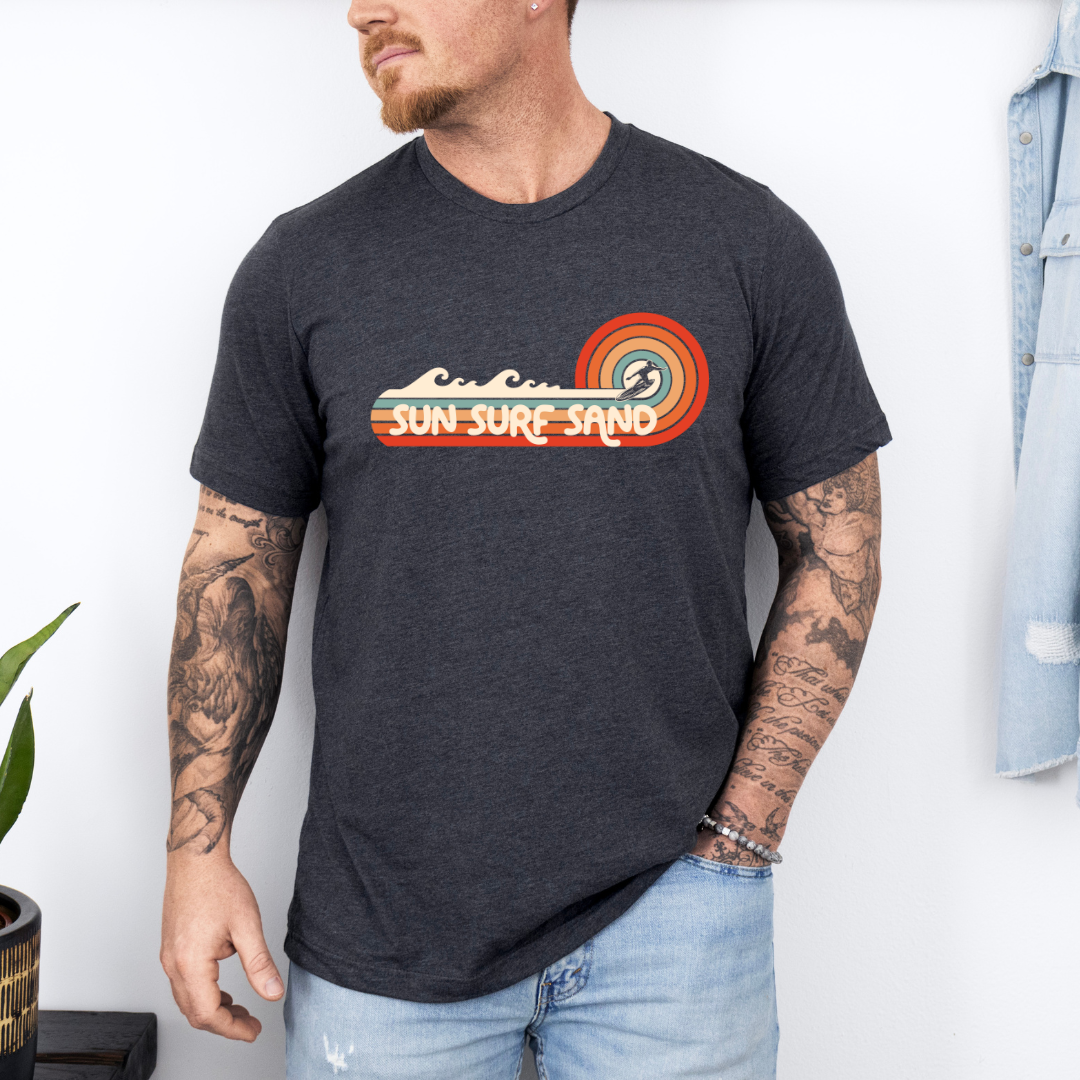 Man wearing a dark gray t-shirt with a retro-style "Sun Surf Sand" graphic design, featuring waves and a sunset, perfect for beach and surf enthusiasts.