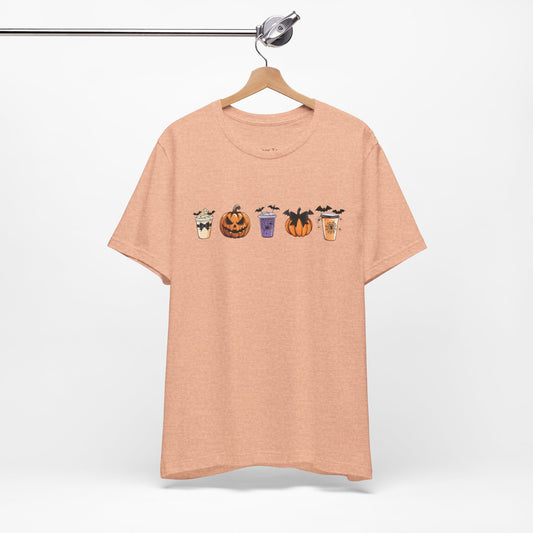 Peach-colored t-shirt featuring five Halloween-themed coffee cups and pumpkins with bats, perfect for coffee lovers and Halloween enthusiasts seeking a fun, spooky vibe.