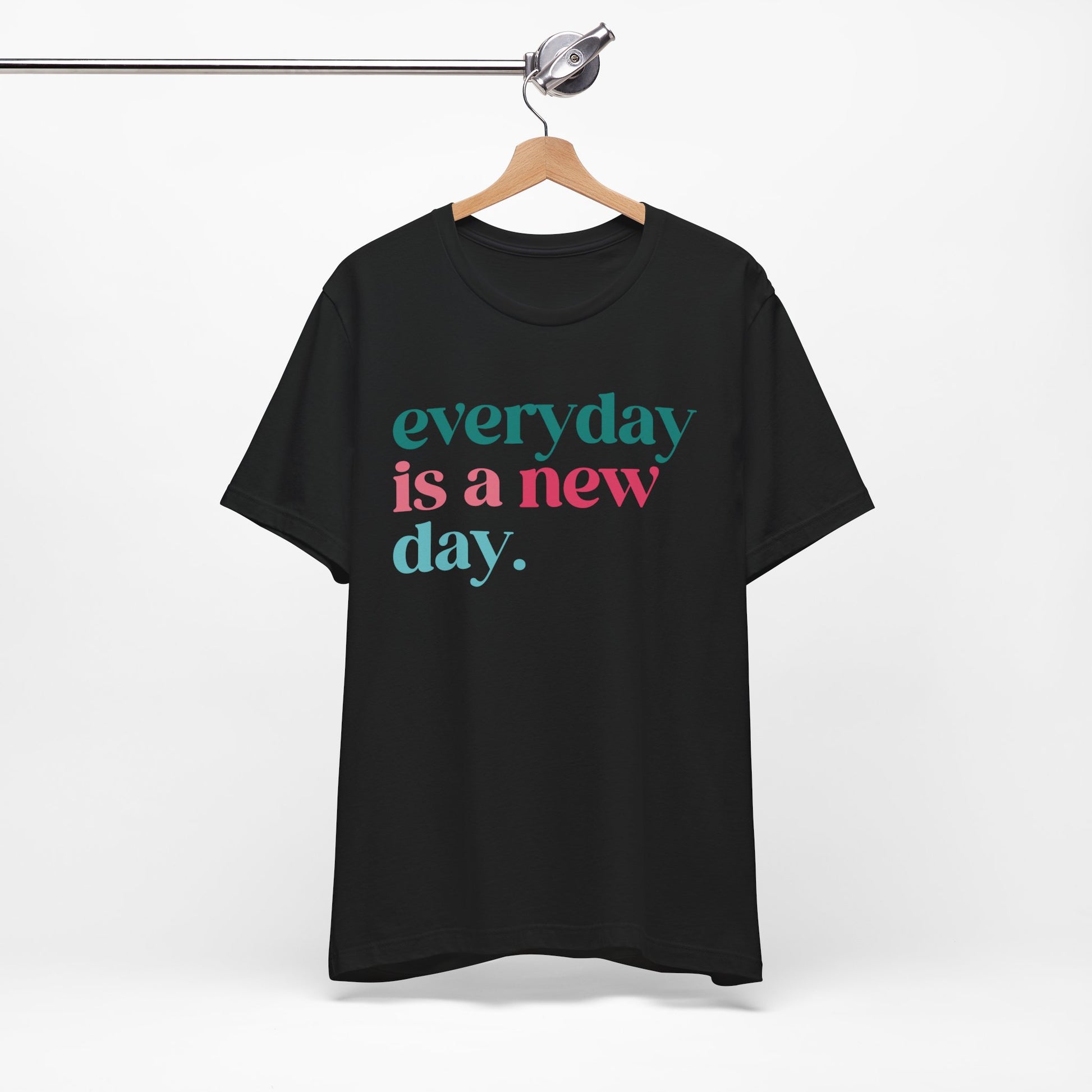 Black t-shirt hanging on a wooden hanger, featuring bold text in colorful fonts that reads, "Everyday is a new day." The design combines teal and pink lettering, offering an uplifting and motivational message. The t-shirt has a relaxed fit and casual style, perfect for daily wear or gifting.