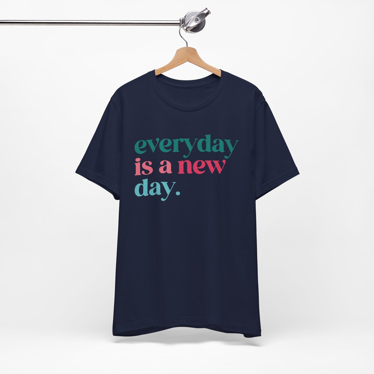 Navy blue t-shirt hanging on a wooden hanger, featuring bold text in colorful fonts that reads, "Everyday is a new day." The design combines teal and pink lettering, offering an uplifting and motivational message. The t-shirt has a relaxed fit and casual style, perfect for daily wear or gifting.